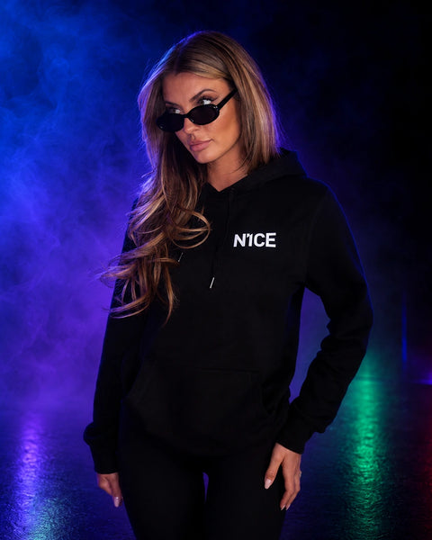 Woman wearing a black N1CE hoodie and sunglasses against a blue smoky backdrop